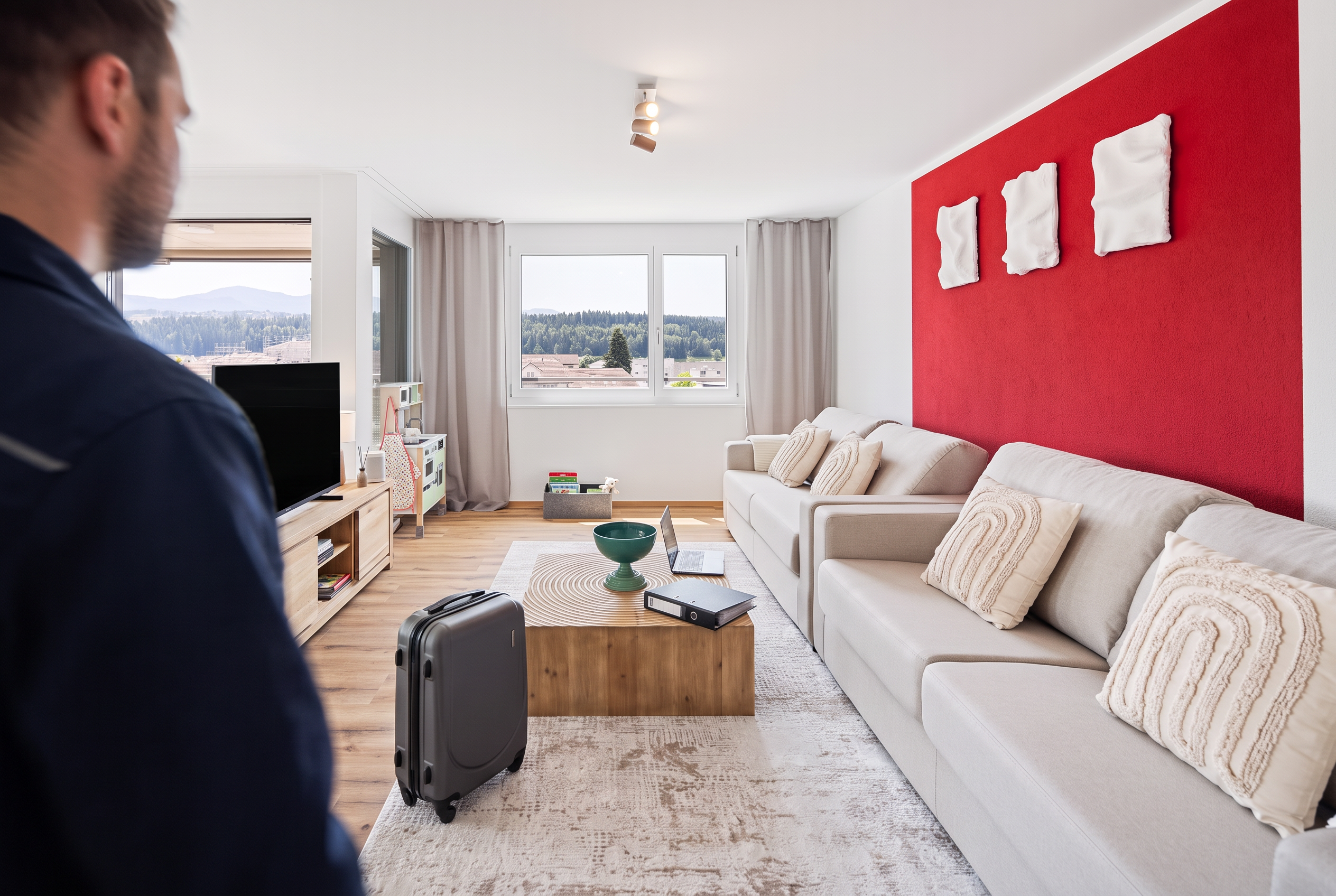 Over-the-Shoulder-Aufnahme: Der Fotograf steht hinter dem Monteur, nur Schulter und Hinterkopf sind im Vordergrund zu sehen, während er in ein helles Schlaf- und Arbeitszimmer mit zwei Betten, Schreibtisch und Koffer auf dem Boden tritt
